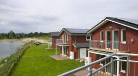 Moderne rote Ferienhäuser mit Balkon und direkter Seelage. Die Häuser haben große Fenster, Solarzellen auf dem Dach und gepflegte Rasenflächen vor dem Wasser