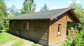 Eingeschossiges Holzferienhaus mit flachem Satteldach und Holzverschalung. Überdachter Eingang, gepflasterter Weg davor, umgeben von Rasen und Bäumen