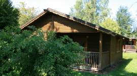 Gemütliches einstöckiges Ferienhaus aus Holz mit Satteldach, überdachter Veranda mit Geländer, teils durch Büsche verdeckt. Umgeben von Bäumen und Rasenfläche