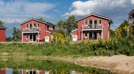 Moderne rote Ferienhäuser mit Balkon und direkter Seelage. Die Häuser haben große Fenster, Solarzellen auf dem Dach und gepflegte Rasenflächen vor dem Wasser