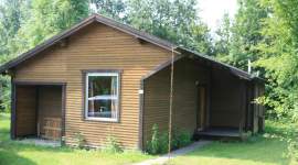 Schönes eingeschossiges Holzferienhaus mit überdachtem Seiteneingang und einem Fenster mit buntem Vorhang, gelegen auf einer Rasenfläche mit Bäumen