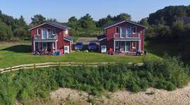 Zwei moderne, rote Ferienhäuser im skandinavischen Stil mit Balkon, großen Fenstern, Holzverkleidung und vorgelagertem Gartenbereich. Davor stehen Autos und Gartenmöbel