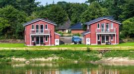 Vier moderne rote Ferienhäuser mit Balkon in direkter Seelage. Gepflegte Rasenflächen, klares Wasser und blauer Himmel im Hintergrund