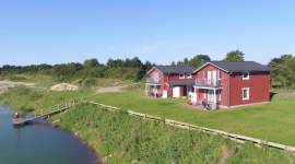 Vier moderne rote Ferienhäuser mit Balkon in direkter Seelage. Gepflegte Rasenflächen, klares Wasser und blauer Himmel im Hintergrund