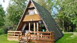 Klassisches A-förmiges Ferienhaus aus Holz mit grünem Schrägdach, kleiner Eingangstreppe und Fenstern im oberen sowie unteren Giebelbereich, umgeben von Bäumen und Wiese