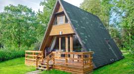 Modernes A-förmiges Holzhaus mit großer Glasfront, Holzterrasse, dunklem Schrägdach und grüner Rasenfläche, umgeben von Bäumen