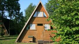 A-förmiges Holzhaus mit steilem Satteldach, mittiger Eingangstür, kleinen Fenstern und umgeben von Bäumen und Rasenfläche