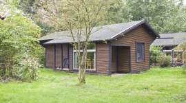 Dunkles Holzferienhaus mit großen Fenstern und überdachter Eingangsveranda, umgeben von Rasenfläche und Bäumen