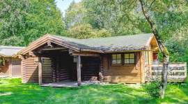 Einstöckiges Blockhaus mit überdachtem Eingangsbereich aus Rundholz und einer Holzveranda rechts. Große Fenster mit Vorhängen, umgeben von Bäumen und Rasenfläche