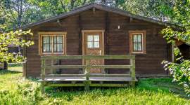 Ein kleines, einstöckiges Holzhaus mit Holzveranda, Fenstern und einer Tür, umgeben von grünem Gras und Bäumen