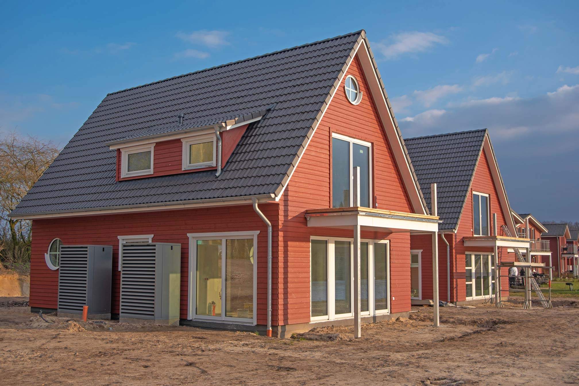 Neues rotes Ferienhaus auf dem Geesthof im Bau, mit grauem Satteldach und großen Fenstern. Weitere baugleiche Häuser im Hintergrund
