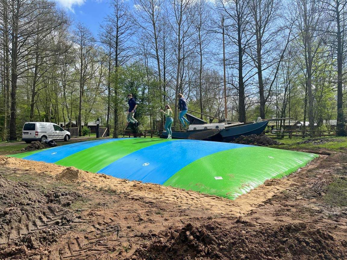 Kinder springen auf einem bunten Luftkissen auf dem Geesthof. Im Hintergrund ein altes Boot und hohe Bäume