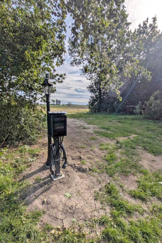 Bild von einer WallBox, die vor einem Durchgang zu Weiden und Felden führt. Der Durchgang ist von Bäumen und Sträuchern begrenzt. Die WallBox Ladesäule für E-Autos steht direkt vor einer schwarzen Weglaterne.