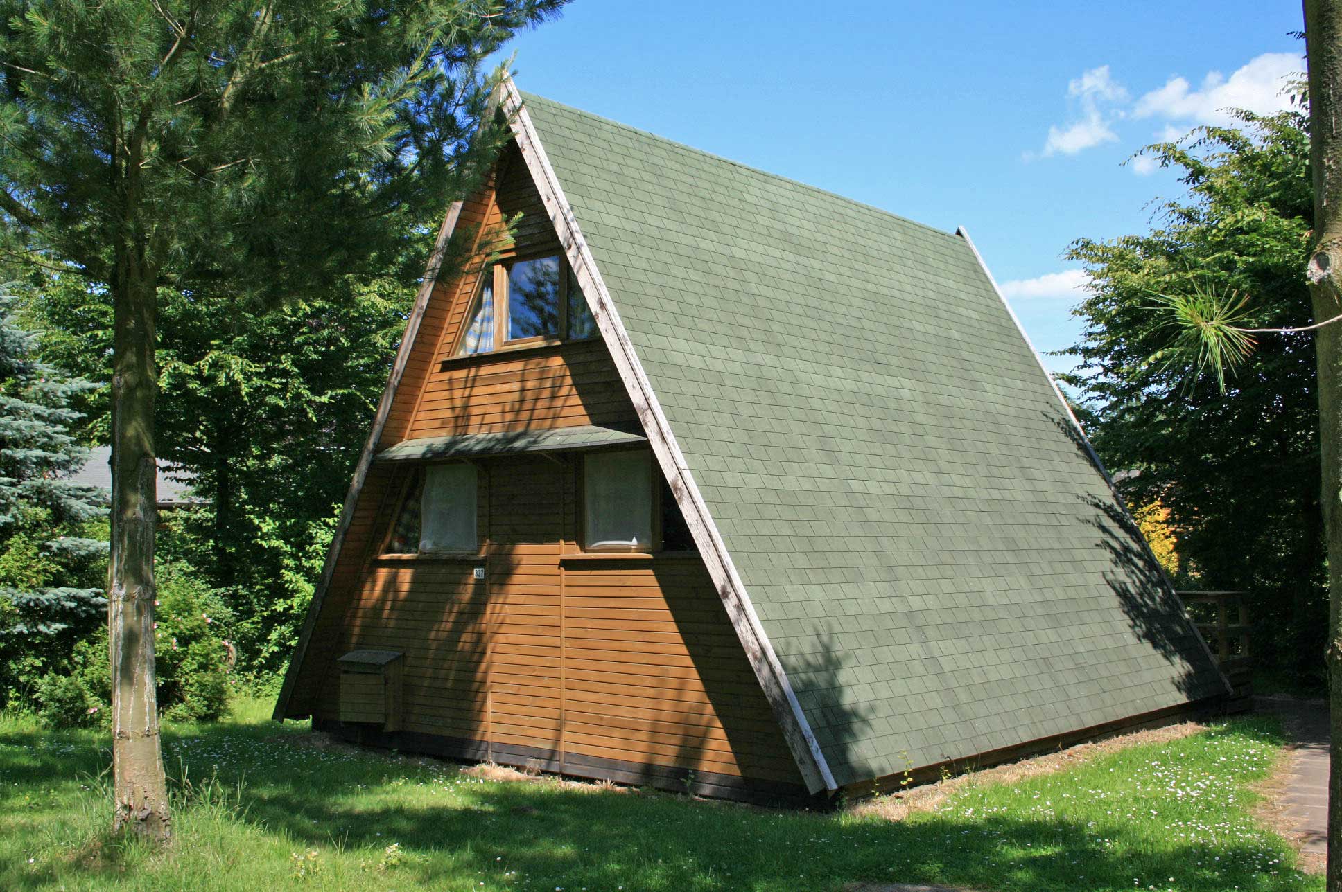 Typ E – Nurdachhaus 337 mit grünem Dach, Holzveranda und Wald im Hintergrund