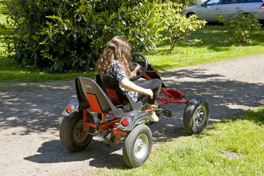 Foto eines Mädchens, das auf einem Kettcar sitzt und einen Schotterweg entlang fährt.