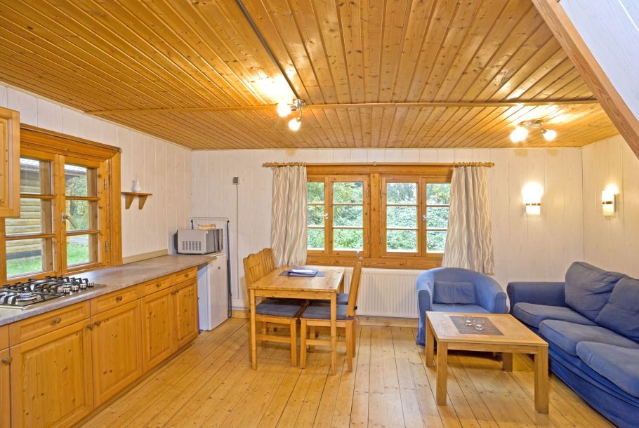 Foto von dem Wohn- und Essbereich im kleinen Blockhaus 325. Auf der linken Seite des Raumes befindet sich die Küchenzeile mit Gaskochfeld, Kühlschrank und Mikrowelle. Mittig im Bild steht ein Esstisch aus Holz an dem vier Holzstühle stehen. Über dem Tisch ist ein Doppelfenster mit Holzrahmen verbaut. Rechts im Zimmer steht ein blaues Sofa mit Polsterkissen, vor dem ein Couchtisch aus Holz steht. Über dem Sofa sind zwei Wandlampen an der Wand montiert.
