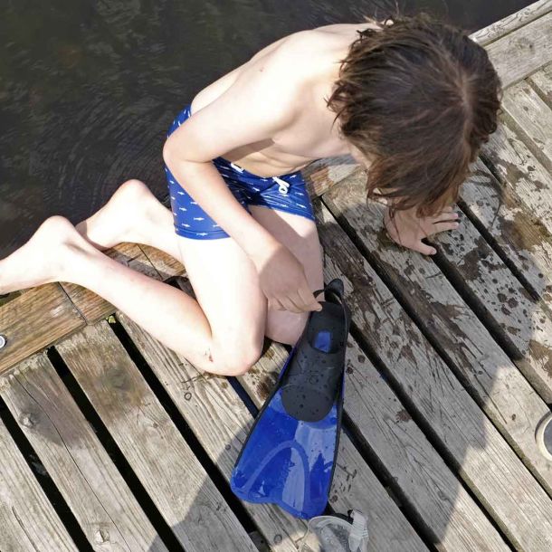 Foto eines Jungen, der mit Taucherflossen auf einem Steg am Wasser sitzt.