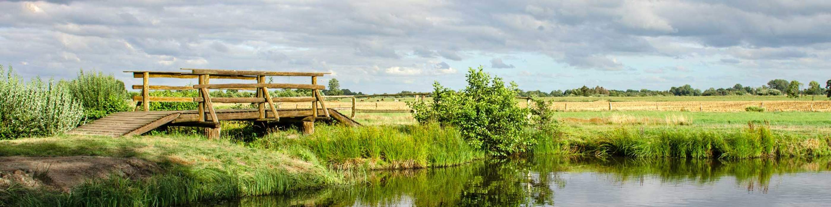 Hölzerne Brücke über einen kleinen Fluss, mit Wiesen und weitem Himmel auf dem Geesthof-Gelände
